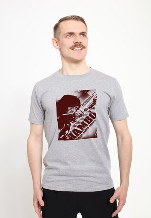 Šedé bavlněné tričko s výrazným červeným grafickým potiskem postavy a nápisem "RAMBO" a "PRVNÍ BLOOD ČÁST II" v retro stylu.
