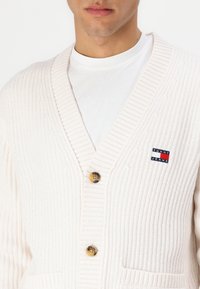 Hvit strikket cardigan med dyp V-hals, to knapper og et lappelogo på venstre side. Teksturt, ribbet stoff med lommer.
