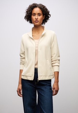 Krämfärgad zip-up jacka med ribbad textur, lättviktig tyg och sidofickor, kombinerad med en åtsittande topp och mörkblå vida jeans.