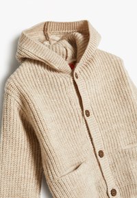 Cardigan lavorato a maglia beige con maniche lunghe, texture a costine, grandi tasche frontali, chiusura con bottoni e un ampio cappuccio ripiegato.