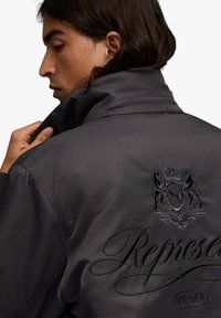Giacca bomber nera con colletto alto, tessuto strutturato, con stemma in rilievo e scritta "Represent" sulla schiena.