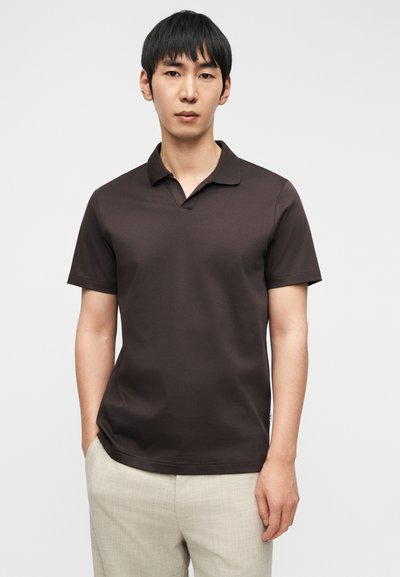 Καφέ κοντομάνικο πόλο με επίπεδη γιακά, κατασκευασμένο από λεία υφάσματα. Διαθέτει στενή γραμμή και ίσια εσοχή, φορεμένο με ανοιχτόχρωμο παντελόνι.