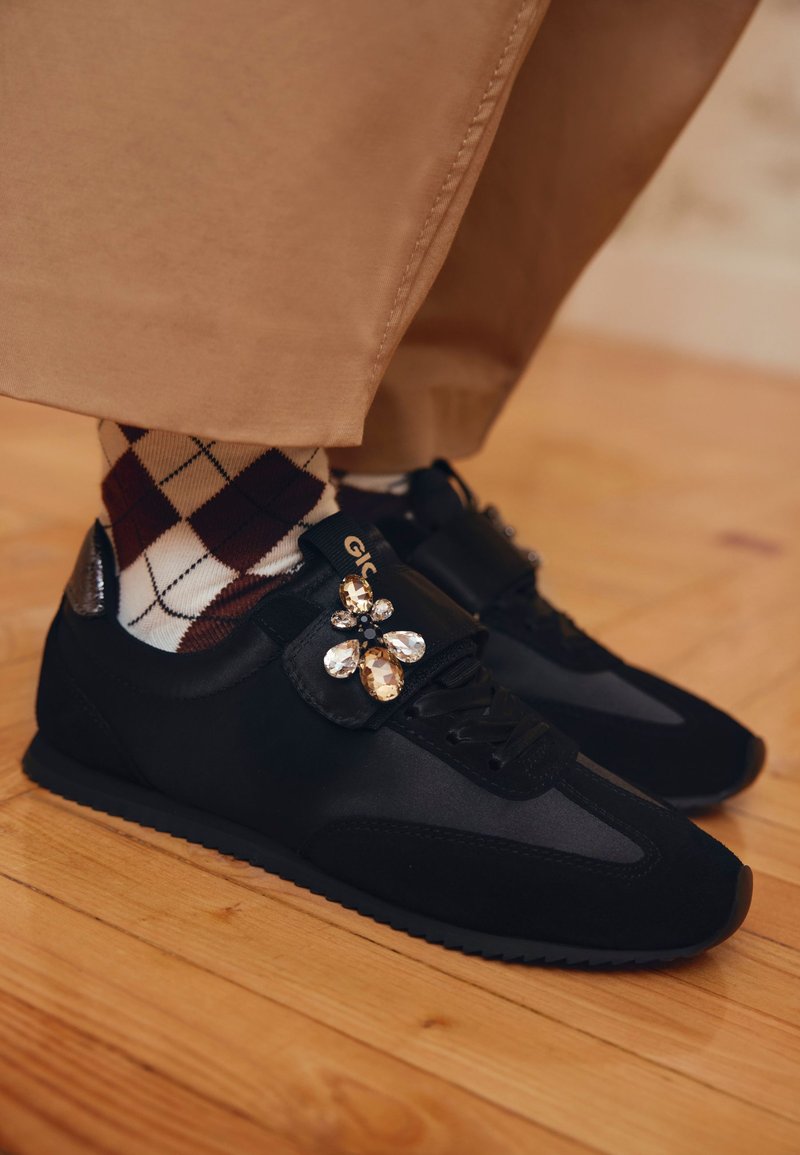 Scarpe atletiche nere con design in materiali misti, tra cui camoscio e raso, con un accent decorativo a forma di gioiello sulla fascia.