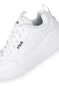 White athletic shoes with a smooth synthetic upper, textured accents, lace-up design, and a cushioned sole. Fila logo in blue and red.