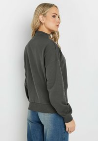 Mörkgrå sweatshirt med hög krage, neddragna axlar och ribbade ärmslut. Bärs tillsammans med ljusblå jeans som visar en avslappnad passform och mjuk textur.