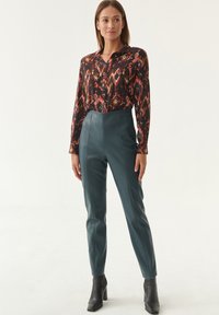 Camicia a maniche lunghe con bottoni, in tessuto nero con motivi geometrici multicolore, abbinata a pantaloni in pelle teal a vita alta e stivaletti neri.
