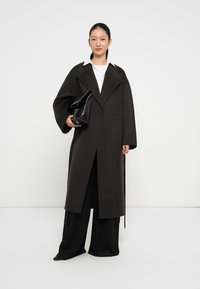 Cappotto lungo color marrone scuro con ampi rever, chiusura doppiopetto e vestibilità morbida. Abbinato a pantaloni neri a gamba larga e una borsa nera.