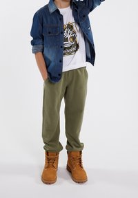 Džínová košile, bílý grafický tričko, olivově zelené tepláky a béžové boty. Obsahuje knoflíky, ohrnuté rukávy a texturované materiály.