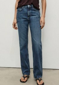 Jeans de mezclilla azul con un diseño de pierna recta, que cuentan con cinco bolsillos, un ligero desgastado y una cintura alta. Se llevan con sandalias negras.
