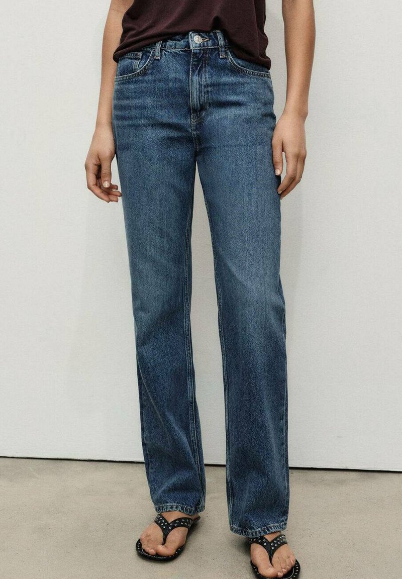 Jeans de mezclilla azul con un diseño de pierna recta, que cuentan con cinco bolsillos, un ligero desgastado y una cintura alta. Se llevan con sandalias negras.