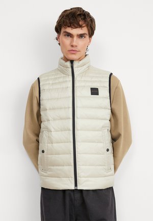 Lichtbeige puffervest met een hoge kraag, ritssluiting aan de voorkant en twee zijzakken. Heeft zwarte zijaccenten en een logopatch op de borst.