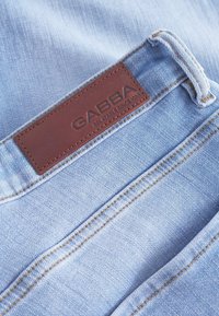 Šviesiai mėlyni džinsai iš džinsinio audinio su rudos odos etikete, ant kurios juosmens juostos įspaustas užrašas „GABBA“. Išsiskiria kontrastingu geltonu siuvimu ir tvirta medžiaga.