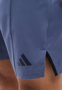 Κοντινή φωτογραφία μπλε σορτς Adidas με μαύρο λογότυπο στο μηρό, που φοριέται από ένα άτομο, δείχνει πλαϊνή σχισμή και λεία υφή υφάσματος.