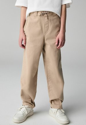 Beige Baumwollhose mit elastischem Bund und Zugbanddetails. Mit Bündchen an den Knöcheln, kombiniert mit weißen Sneakern.