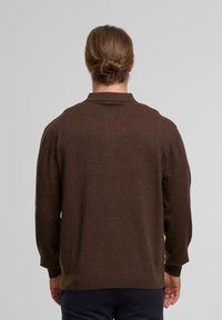 Person mit braunen Haaren im Dutt, trägt einen braunen Langarmpullover und dunkle Hose, von hinten vor einem schlichten Hintergrund gesehen.