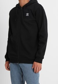 Man med svart Adidas zip-up hoodie med huva och ljusblå jeans, som står mot en helt vit bakgrund.