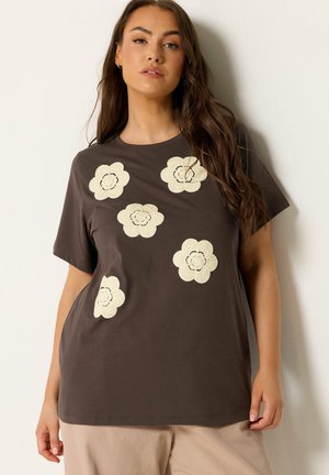 Kvinde med langt hår iført en mørkebrun t-shirt med fem beige blomsterdesigns, stående mod en lys baggrund.