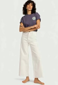 Grafiska T-kreklis purpursarkanā krāsā ar apļveida zilu dizainu, savienots ar augsto jostasvietu baltām platām biksēm. Audums izskatās mīksts ar saplēstiem malām.