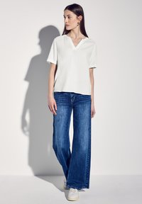 Blusa bianca a maniche corte con scollo a V, abbinata a jeans in denim blu a gamba larga. Il modello indossa sneakers bianche. Tessuto morbido, vestibilità comoda.