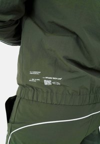 Groene nylon jas met een gestructureerd oppervlak, uitgerust met witte piping en een label met merknaam en QR-code. Zichtbaar detail van een elastische tailleband.