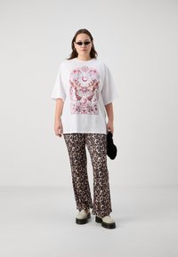 Bílé oversized tričko s grafikou motýla, doplněné kalhotami se zvěří v kontrastním flare střihu. Model má na sobě černé sluneční brýle a bílé platformové boty.