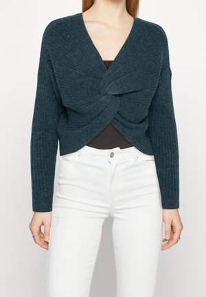 Femme portant un pull en maille bleu sarcelle foncé torsadé à l'avant sur un top noir, associé à un jean blanc taille haute sur un fond uni.