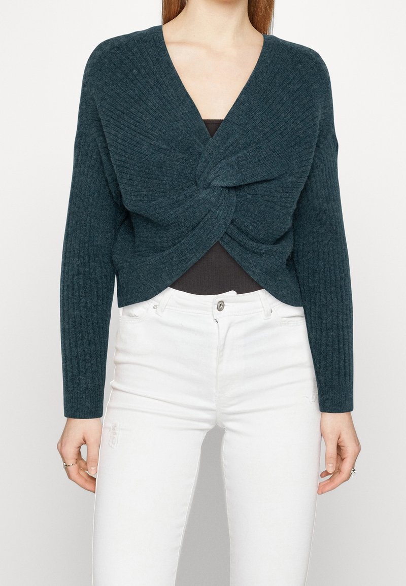 Femme portant un pull en maille bleu sarcelle foncé torsadé à l'avant sur un top noir, associé à un jean blanc taille haute sur un fond uni.