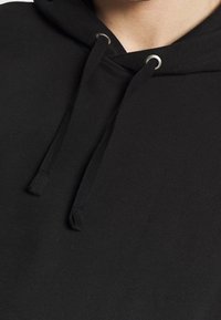 Gros plan sur l'encolure d'un sweat à capuche noir avec des cordons passés dans des œillets en métal, au niveau du cou et des épaules d'une personne.