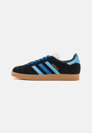 Černé semišové tenisky s modrými akcenty, třemi modrými pruhy, gumovou podrážkou a bílým jazykem. Na boku zlaté logo "Gazelle".