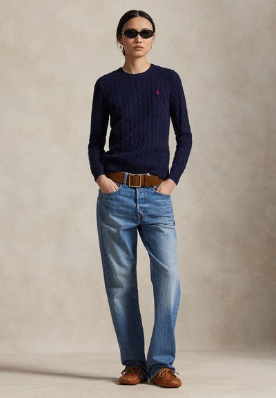 Femeie purtând ochelari de soare negri, pulover albastru închis cu model torsadat, curea maro, blugi albaștri largi și mocasini maro, stând cu mâinile în buzunare.