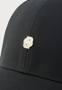 Șapcă neagră din material texturat, cu un emblemă aurie „B” pe partea din față. Design simplu, cu o formă clasică și finisaj neted.