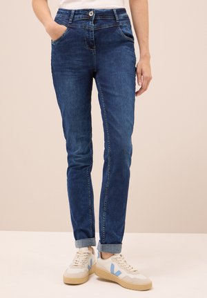 Indigo spijkerbroek met een slim fit, voorzien van voorzakken, een opgerolde zoom en een knoopsluiting. Gecombineerd met witte sneakers.
