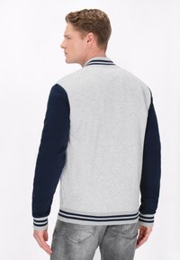 Jachetă din bumbac gri cu mâneci bleumarin, guler ribionat și manșete stripe. Prezintă o textură netedă și o croială lejeră.