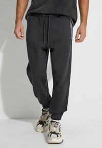 Männliche dunkelgraue Jogginghosen aus einem weichen, strukturierten Stoff mit elastischem Bund, Kordelzug und rippgestrickten Knöcheln. Kombiniert mit beigen Sneakers.