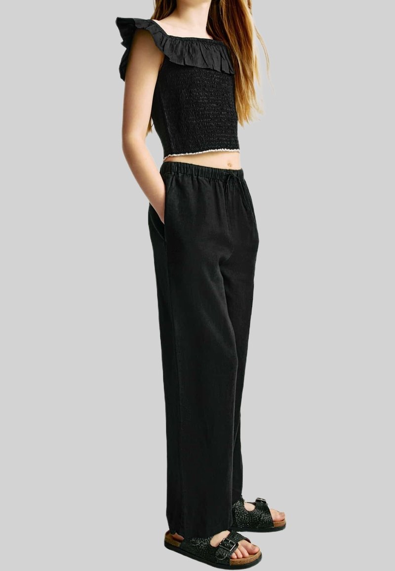 Fille portant un crop top noir à volants et épaules dénudées, un pantalon noir ample avec poches et des sandales noires à boucles, debout les mains dans les poches.