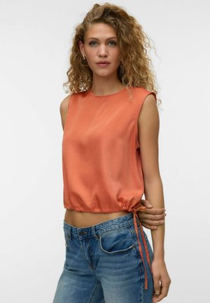 Kvinde med krøllet hår iført en ærmeløs orange top med snøre og blå jeans, står mod en ensartet hvid baggrund.