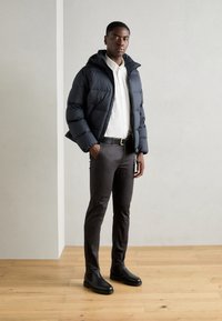 Jeune homme debout à l'intérieur portant une veste matelassée noire, une chemise blanche à boutons, un pantalon sombre, une ceinture noire et des bottes noires sur un sol en bois.
