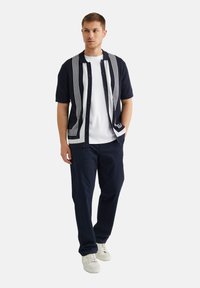 Navy short-sleeve cardigan with white vertical stripes, button closure, worn over a white t-shirt, paired with dark pants and white sneakers.