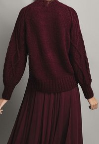 Burgundy neulepaita, jossa on kaapelikuvioita hihoissa, ribattu helma ja rento muotoilu, yhdistettynä samaan sävyyn pitkän hameen kanssa.