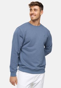 Suéter azul de suave tejido, con cuello redondo y puños acanalados. Logo sutil en el lado izquierdo del pecho. Se lleva con pantalones blancos.