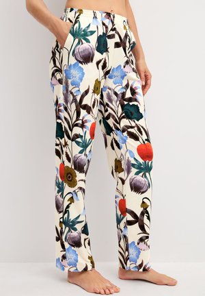 Vrouw draagt losse crèmekleurige broek met grote kleurrijke bloemmotieven, staat blootsvoets met één hand in de zak tegen een effen achtergrond.