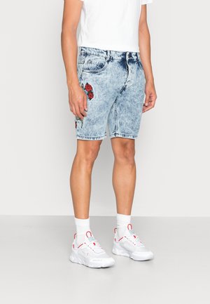 Osoba nosząca krótkie spodenki z denimu w stylu acid wash z czerwonym haftem róży, białe skarpetki, białe i czerwone trampki oraz gładką białą koszulkę.