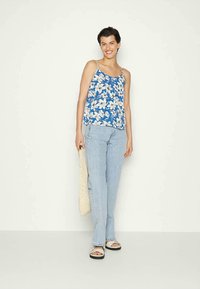Camisola sem mangas azul floral com flores brancas, jeans azul claro e sandálias pretas. Modelo segura uma bolsa tecida bege, em pé contra um fundo simples.