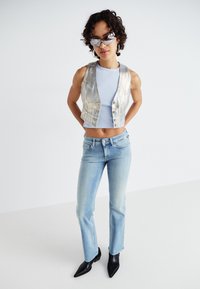 Metaalachtige zilveren cropped vest, lichtblauw aansluitend tanktop, lichtblauwe jeans en puntige zwarte enkellaarsjes. Strakke, reflecterende texturen en moderne pasvorm.