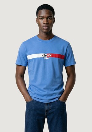 Joven con una camiseta azul Tommy Jeans con rayas blancas y rojas y vaqueros azul oscuro, de pie con las manos en los bolsillos.