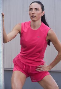 Atletický outfit v jasně růžové barvě, který se skládá z upnutého topu a volných šortek s mřížkovaným vzorem. Na topu je logo Nike. Hladká textura látky.