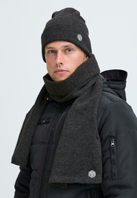 Nuori mies, jolla on päässään tummanharmaa neulepipo ja siihen sopiva huivi, mustan tikatun takin kanssa vaaleata taustaa vasten.