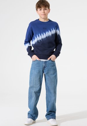 Suéter de punto con estampado de teñido anudado en azul marino y acentos en blanco, con cuello redondo y puños acanalados, combinado con unos vaqueros de corte holgado en azul claro y zapatillas blancas.