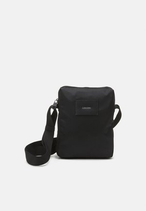 Kompakt sort crossbody taske med justerbar rem og Calvin Klein logo patch foran, ensfarvet baggrund.