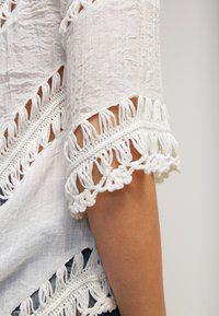 Une manche de vêtement en dentelle blanche avec des détails en crochet et des franges élaborés sur le bras supérieur d'une personne, sur un fond uni.